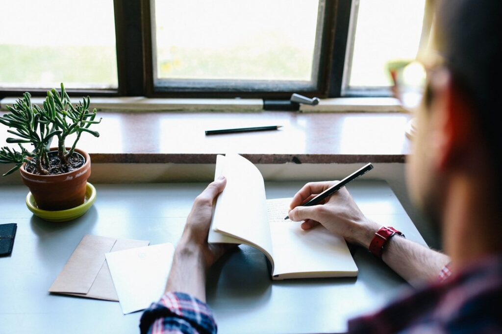 A person writing in a journal at a desk by the window